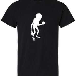 May include: Black t-shirt featuring a white silhouette of a creature holding a ball. The design is centered on the front of the short-sleeved shirt. The creature has a furry appearance with a round head and a ball in its hand.