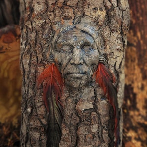 Może przedstawiać: Rzeźba twarzy z zamkniętymi oczami, wyrzeźbiona w drewnie i osadzona w pniu drzewa. Rzeźba jest pomalowana w odcieniach brązu, szarości i czerwieni. Po obu stronach twarzy znajdują się dwie czerwone pióra.