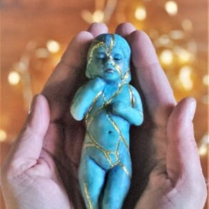 Może przedstawiać: Mała, niebieska figurka przedstawiająca osobę z złotymi akcentami. Figurka jest trzymana w dłoniach.
