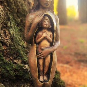 May include: A bronze-toned sculpture of a woman holding a smaller figure within her torso. The sculpture is set against a natural backdrop of a tree trunk and blurred foliage. The figures are detailed, with a focus on the textures and forms.