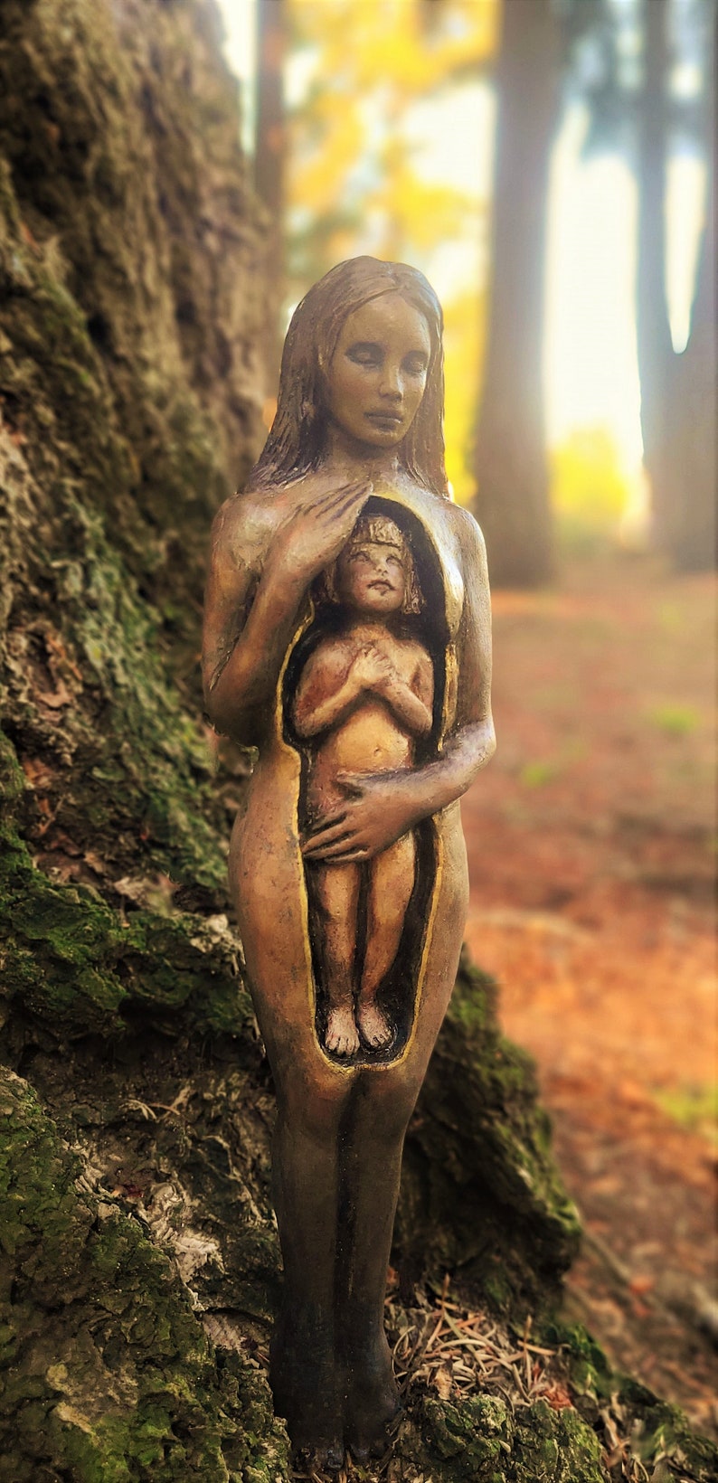 May include: A bronze-toned sculpture of a woman holding a smaller figure within her torso. The sculpture is set against a natural backdrop of a tree trunk and blurred foliage. The figures are detailed, with a focus on the textures and forms.