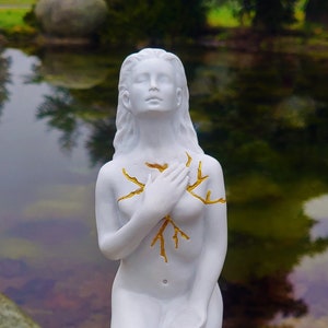 May include: White statue of a person kneeling with their hands clasped over their chest. The statue has gold painted lines on the chest area.