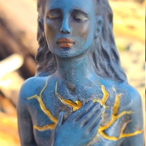 May include: A blue statue of a woman with her hands clasped over her heart. The statue has gold accents on the chest and hair.