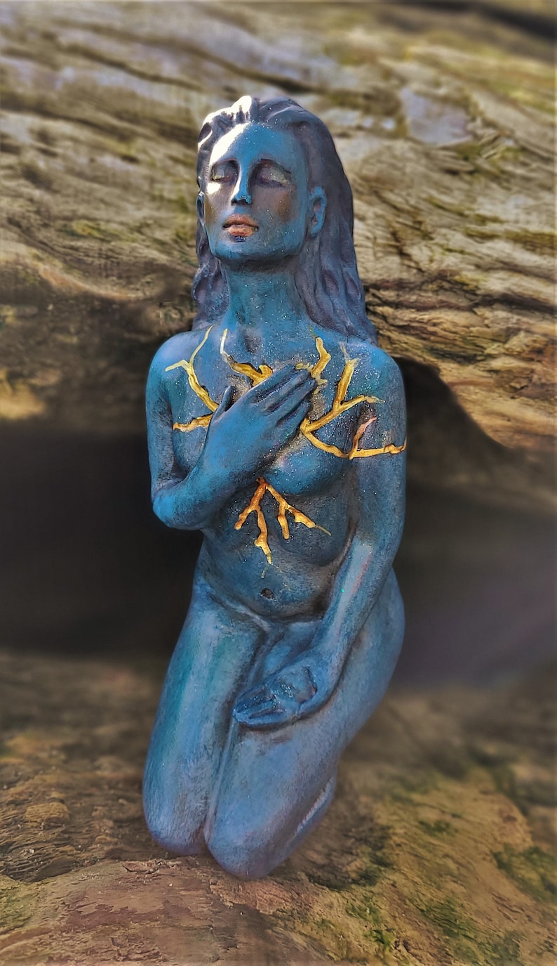 May include: A blue ceramic sculpture of a woman kneeling with her hands clasped in front of her chest. The sculpture has gold paint details on the chest area, resembling branches or roots.