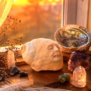 May include: A beige seashell sculpture carved with a human face featuring closed eyes and a serene expression, positioned alongside an iridescent abalone shell, several raw amethyst and clear quartz crystals, a small green stone, and a white feather.