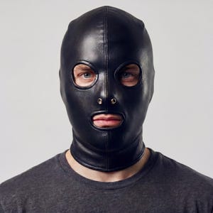 May include: Black leather hood with eye and mouth openings. The hood has a vertical seam down the center and a small zipper detail at the nose. The neck area is fitted. The person is wearing a gray t-shirt.