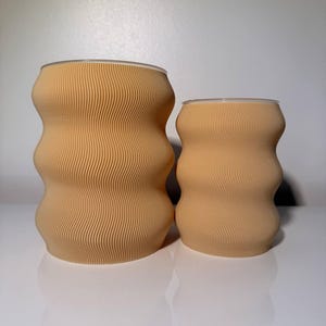 May include: Two beige-colored, wavy-textured drinking glasses. The glasses have a unique, hourglass-like shape with a ridged surface. The rims of the glasses are clear. The glasses are of different sizes, with one slightly larger than the other.