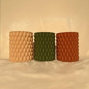May include: Three cylindrical planters in a row, each with a textured, repeating diamond pattern. The planters are in shades of tan, olive green, and reddish-brown. The background is a solid, light beige color.