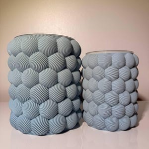 May include: Two cylindrical containers in a light blue color, featuring a textured exterior composed of numerous small, spherical protrusions with a fine, diagonal ribbed pattern covering the entire surface of both objects.