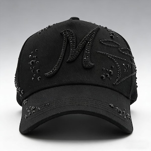May include: Black baseball cap with a curved brim. The cap features a large, embellished letter "M" on the front, along with scattered decorative accents. The cap is made of a textured fabric.