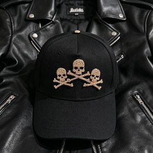 May include: Black baseball cap adorned with three rhinestone skull and crossbones motifs. The cap is displayed on a black leather jacket, offering a striking contrast. The design of the cap gives a rock and roll vibe.