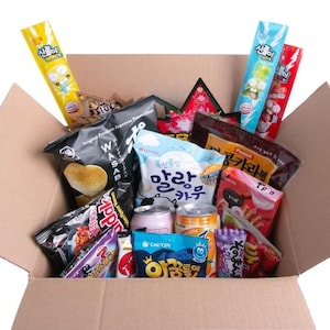 May include: A cardboard box overflowing with a selection of Asian snacks. The box is packed with colourful packets of crisps, sweets, and drinks. Some packets feature Korean and Japanese text. The snacks are arranged inside the open box.