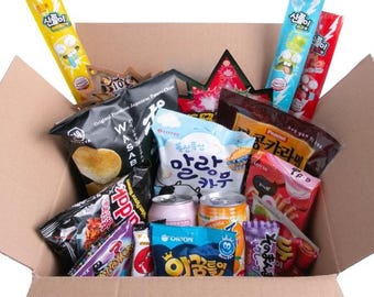May include: A cardboard box filled with various Asian snacks. The box contains colorful packages of chips, candies, and drinks. Some packages have Korean and Japanese text. The snacks are arranged inside the open box.