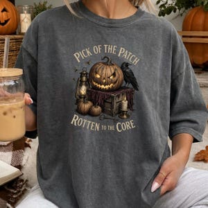May include: A charcoal gray t-shirt featuring a graphic print of a glowing jack-o'-lantern, a black raven, a lantern, and a candle on a wooden crate. The text reads PICK OF THE PATCH ROTTEN TO THE CORE in a beige font, centered on the chest.