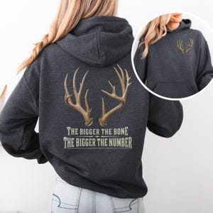 May include: A dark charcoal gray hooded sweatshirt featuring a large graphic on the back of two deer antlers with the text "THE BIGGER THE BONE THE BIGGER THE NUMBER" printed below. A smaller version of the antler graphic is on the front left chest.