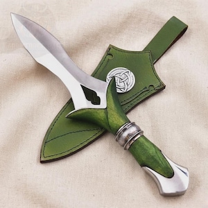 Puede incluir: Una daga artesanal con una hoja de acero pulido y un mango de madera verde. El mango está adornado con anillos de metal plateado y un extremo decorativo. Viene con una funda de cuero verde con un emblema de nudo celta.
