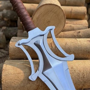 May include: A polished silver sword with a brown leather-wrapped handle. The blade has a distinctive shape with a central ridge and curved guard. The sword rests on a background of stacked wooden logs. The text "NORDICFORGECRAFTS" is visible.