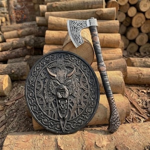 May include: A circular shield featuring a central relief of a snarling wolf head surrounded by intricate knotwork patterns, paired with a battle axe that has a silver-toned blade engraved with Celtic-style designs and a dark brown leather-wrapped handle.