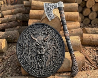 May include: A circular shield featuring a central relief of a snarling wolf head surrounded by intricate knotwork patterns, paired with a battle axe that has a silver-toned blade engraved with Celtic-style designs and a dark brown leather-wrapped handle.