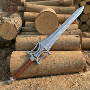 May include: A silver-colored sword with a brown leather-wrapped handle. The blade is long and pointed, with a decorative hilt. The sword is displayed on a bed of wooden logs. The text "NORDICFORGEFACTS" is visible in the upper left corner.