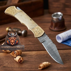 May include: A folding knife with a patterned blade and a handle featuring gold-colored metal accents and a cream-colored grip. The knife is displayed on a wooden surface with wood shavings and the "Nordic Forge Crafts" logo.