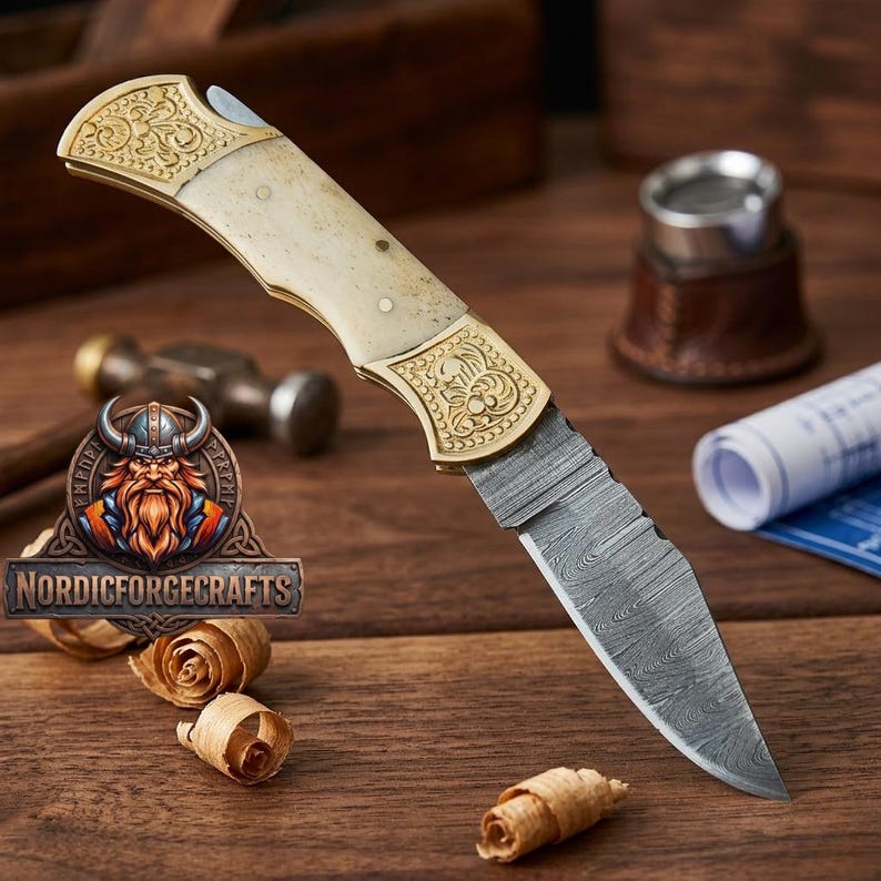 May include: A folding knife with a patterned blade and a handle featuring gold-colored metal accents and a cream-colored grip. The knife is displayed on a wooden surface with wood shavings and the "Nordic Forge Crafts" logo.