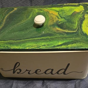 Könnte beinhalten: Rechteckige Brotdose mit cremefarbenem Boden und Deckel mit grün-gelbem Wirbelmuster. Der Deckel hat einen weißen Knopf. Das Wort "bread" ist in Schreibschrift auf die Vorderseite geschrieben.