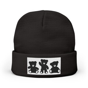 Puede incluir: Gorro negro con un parche rectangular blanco con tres siluetas de osos de peluche negros. Los osos están en diferentes poses, uno sentado, uno saludando y otro sentado.