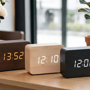 Pode incluir: Três despertadores digitais de madeira em cores diferentes: castanho escuro, madeira clara e preto. Cada relógio exibe a hora em formato digital. Os relógios são retangulares com cantos arredondados e estão em uma superfície de madeira.