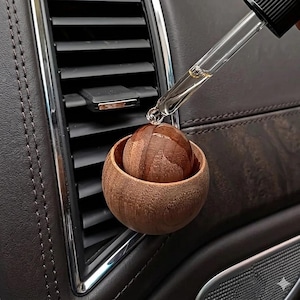 Könnte beinhalten: Ein hölzerner Auto-Lufterfrischer mit kugelförmigem Design. Eine Glaspipette gibt Flüssigkeit auf die Holzkugel ab. Der Lufterfrischer befindet sich in einer Lüftungsöffnung im Auto, mit einem dunkelbraunen Lederinterieur im Hintergrund.