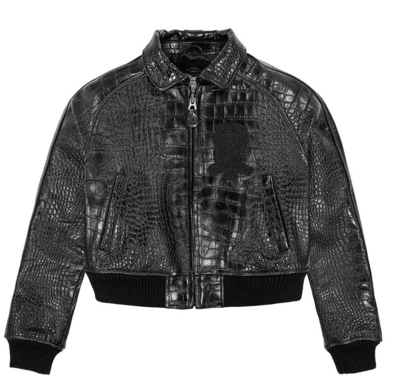May include: A black bomber jacket featuring a textured crocodile skin pattern throughout. The jacket includes a front zipper closure, a standard collar, two side pockets, and black ribbed knit cuffs and hem. A dark patch is visible on the chest.