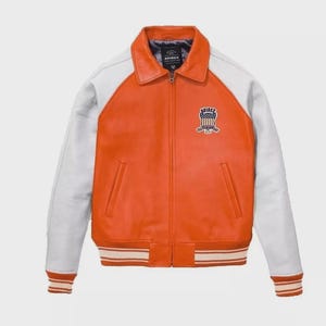 Puede incluir: Una chaqueta de cuero naranja y blanca con cremallera frontal completa, cuello cl&aacute;sico y pu&ntilde;os y dobladillo acanalados. El pecho izquierdo presenta un parche bordado con el texto AVIREX y un gr&aacute;fico de escudo con estrellas y rayas.