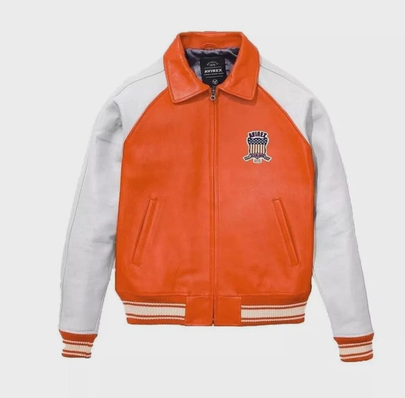 Puede incluir: Una chaqueta de cuero naranja y blanca con cremallera frontal completa, cuello cl&aacute;sico y pu&ntilde;os y dobladillo acanalados. El pecho izquierdo presenta un parche bordado con el texto AVIREX y un gr&aacute;fico de escudo con estrellas y rayas.