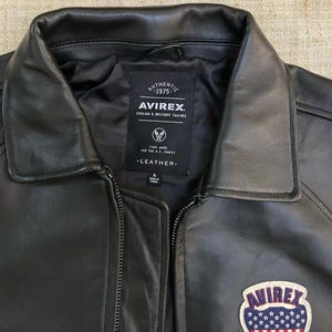 May include: Black leather jacket with a zip closure and a classic collar. The jacket features a patch with the text "AVIREX" and an American flag design. The jacket also has a label with the text "AUTHENTIC 1975" and "AVIREX CIVILIAN & MILITARY TAILORS".