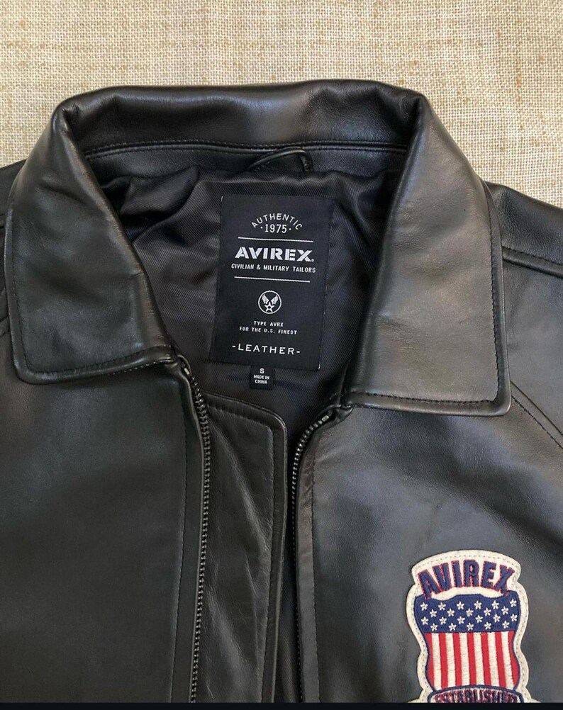 May include: Black leather jacket with a zip closure and a classic collar. The jacket features a patch with the text "AVIREX" and an American flag design. The jacket also has a label with the text "AUTHENTIC 1975" and "AVIREX CIVILIAN & MILITARY TAILORS".