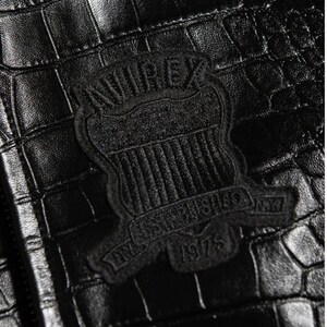 May include: A black jacket with a textured crocodile skin pattern. A black embroidered patch is centered on the material, featuring the text AVIREX at the top, a shield design in the middle, and the text ESTABLISHED 1975 at the bottom.