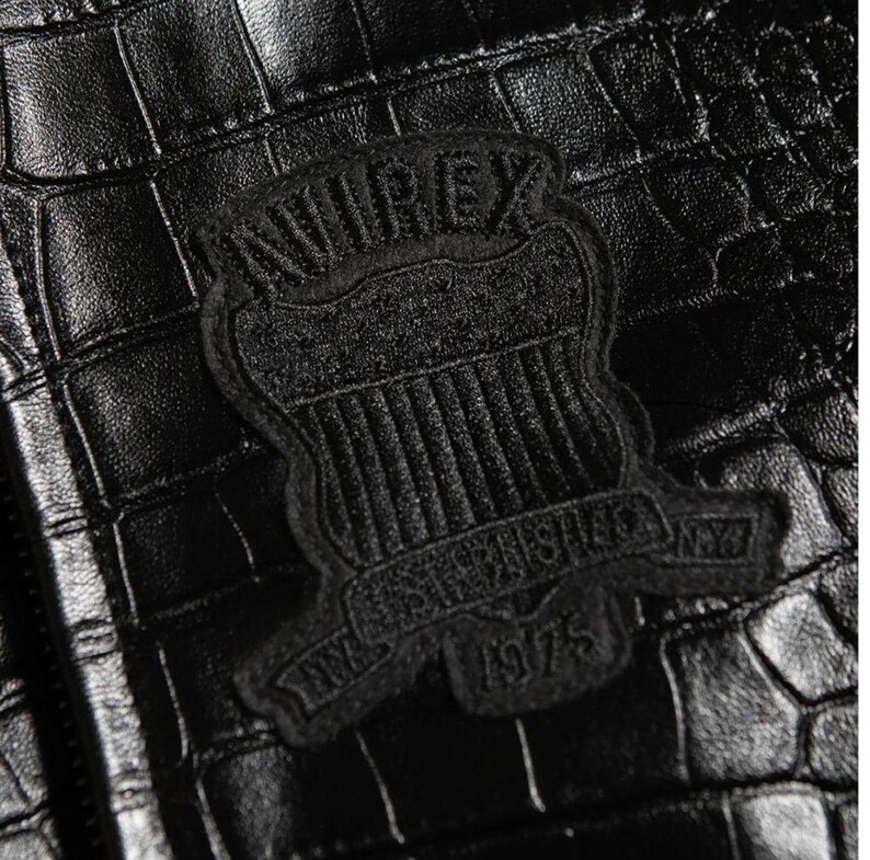May include: A black jacket with a textured crocodile skin pattern. A black embroidered patch is centered on the material, featuring the text AVIREX at the top, a shield design in the middle, and the text ESTABLISHED 1975 at the bottom.