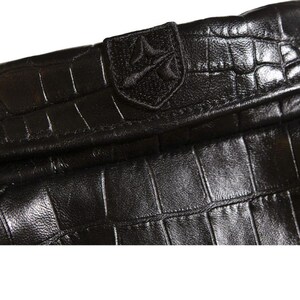 May include: A black leather accessory featuring a prominent crocodile skin texture and a glossy finish. A small, rectangular patch is stitched onto the top edge, displaying an embroidered emblem with a stylized geometric design in black thread.