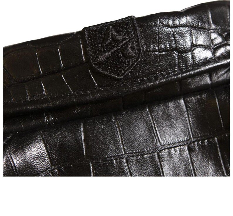 May include: A black leather accessory featuring a prominent crocodile skin texture and a glossy finish. A small, rectangular patch is stitched onto the top edge, displaying an embroidered emblem with a stylized geometric design in black thread.