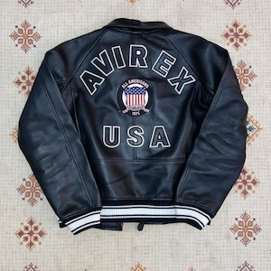 May include: Black leather jacket featuring the word "AVIREX" in white block letters across the back. Below is a patch with "ALL AMERICANS" and "USA" in white. The jacket has a white and black striped trim at the waist and cuffs.