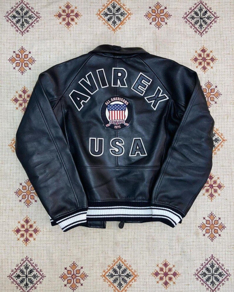 May include: Black leather jacket featuring the word "AVIREX" in white block letters across the back. Below is a patch with "ALL AMERICANS" and "USA" in white. The jacket has a white and black striped trim at the waist and cuffs.