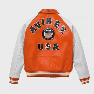 Puede incluir: Una chaqueta universitaria de cuero naranja y blanca con el texto AVIREX en letras negras en la parte superior de la espalda, un parche circular con un gr&aacute;fico de bandera y el texto ALL AMERICANS ESTABLISHED 1975, y la palabra USA debajo.