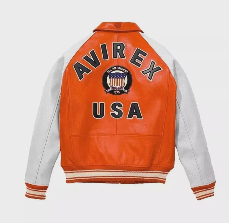 Puede incluir: Una chaqueta universitaria de cuero naranja y blanca con el texto AVIREX en letras negras en la parte superior de la espalda, un parche circular con un gr&aacute;fico de bandera y el texto ALL AMERICANS ESTABLISHED 1975, y la palabra USA debajo.