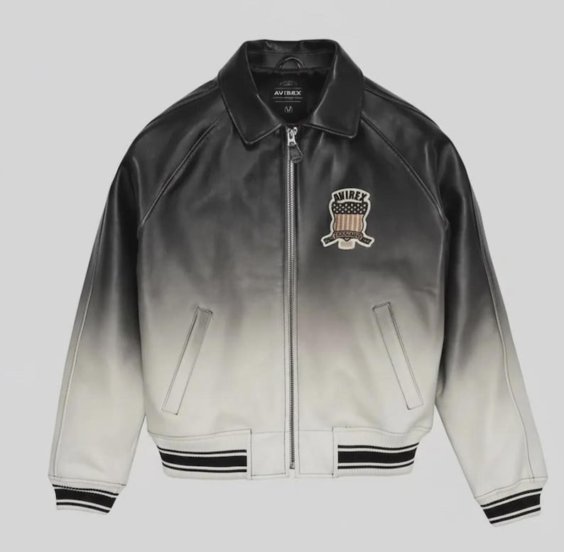 May include: A black and white ombre leather jacket featuring a full-length front zipper, a classic collar, and ribbed cuffs and hem with black and white stripes. An embroidered patch on the left chest displays the text AVIREX above a shield graphic.