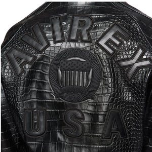 May include: A black jacket with a textured crocodile-skin pattern. The back features large black block letters spelling AVIREX and USA. A central circular patch displays a shield emblem with the text ALL AMERICAN and ESTABLISHED 1975.