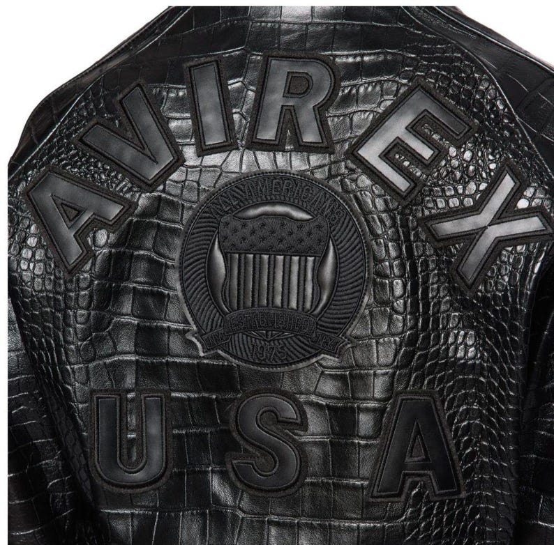 May include: A black jacket with a textured crocodile-skin pattern. The back features large black block letters spelling AVIREX and USA. A central circular patch displays a shield emblem with the text ALL AMERICAN and ESTABLISHED 1975.