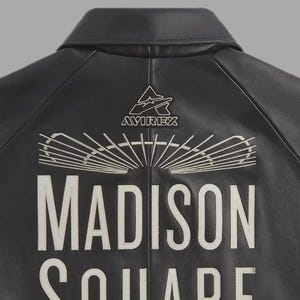 May include: A black leather jacket featuring white embroidered text that reads MADISON SQUARE. Above the text is a white embroidered graphic of a stadium roof and the Avirex logo. The jacket has a classic collar and visible seam detailing.
