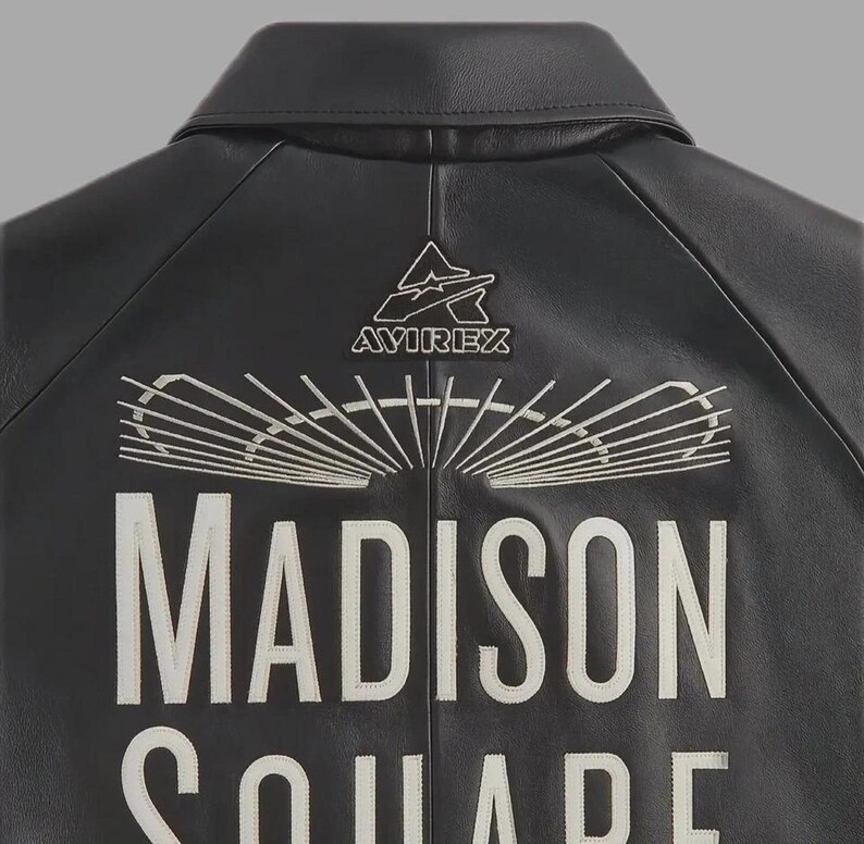 May include: A black leather jacket featuring white embroidered text that reads MADISON SQUARE. Above the text is a white embroidered graphic of a stadium roof and the Avirex logo. The jacket has a classic collar and visible seam detailing.