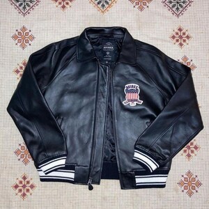 May include: Black leather jacket with a zip-up front, collar, and ribbed cuffs and hem. The jacket features a patch on the chest with the text "AVIREX" and a design of red and white stripes and blue accents.