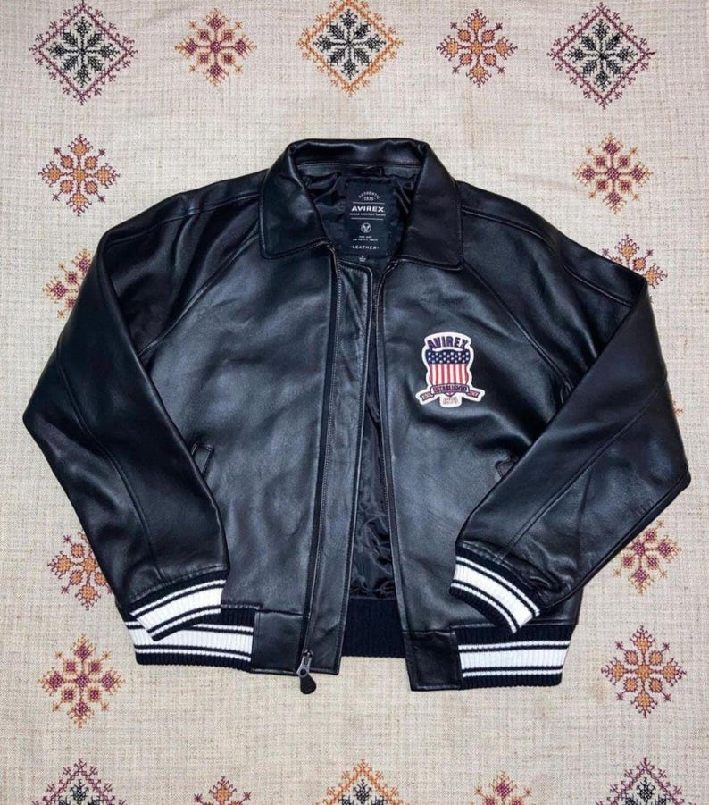 May include: Black leather jacket with a zip-up front, collar, and ribbed cuffs and hem. The jacket features a patch on the chest with the text "AVIREX" and a design of red and white stripes and blue accents.